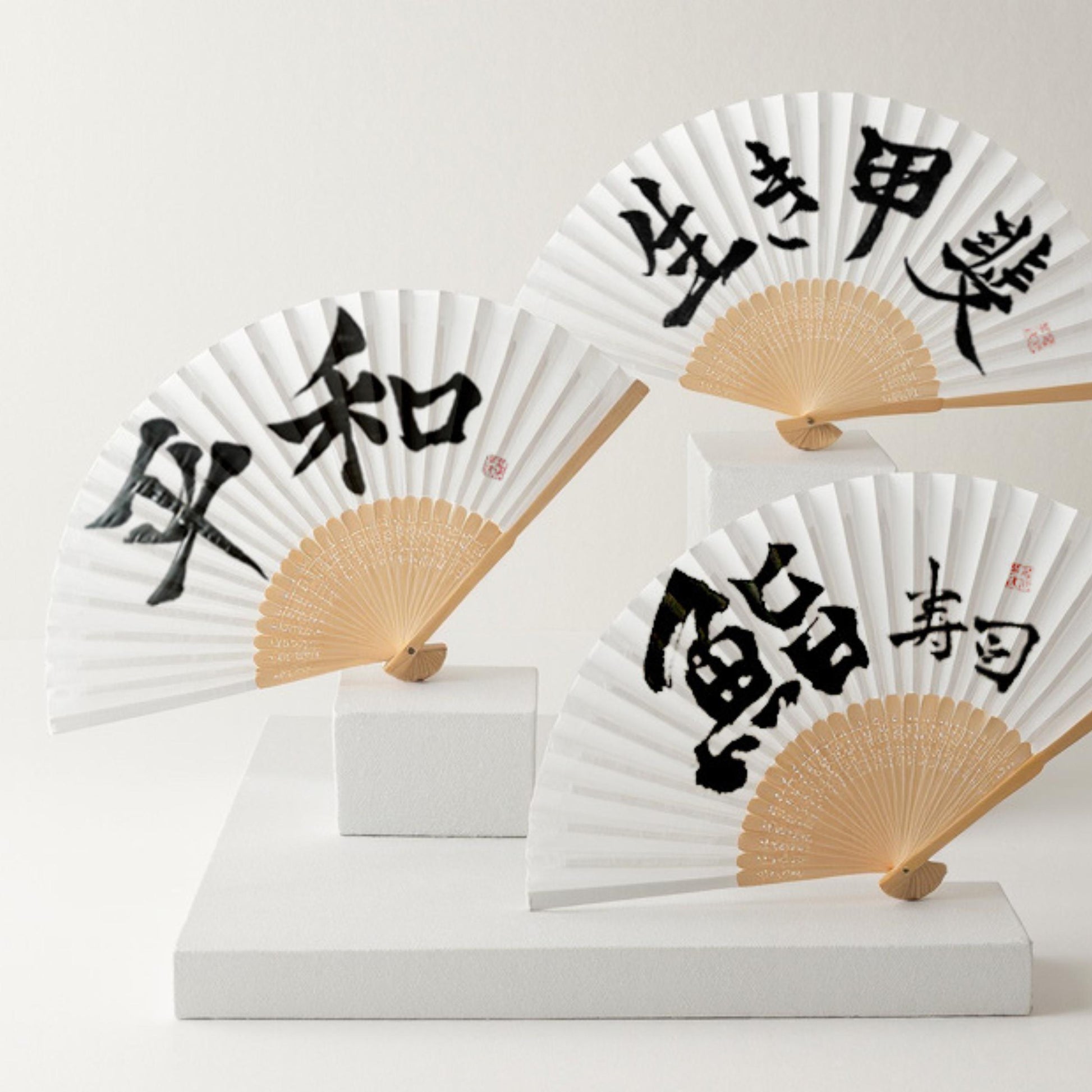 Three folding fans with black Japanese text on white paper, displayed on white stands against a light gray background.
