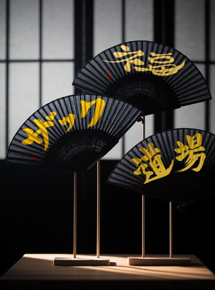 Three black fans with gold kanji on a wooden stand against a dark background