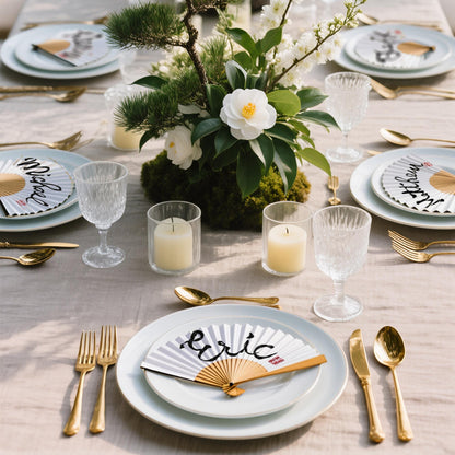 Japanese-style table setting featuring white and gold hand-written folding fans as place cards with the names 'Eric,' 'Michael,' and 'Matthew.' Accompanied by a central arrangement of pine branches, white flowers, moss, and burning candles. Perfect for an elegant Japandi dinner party or wedding decor.