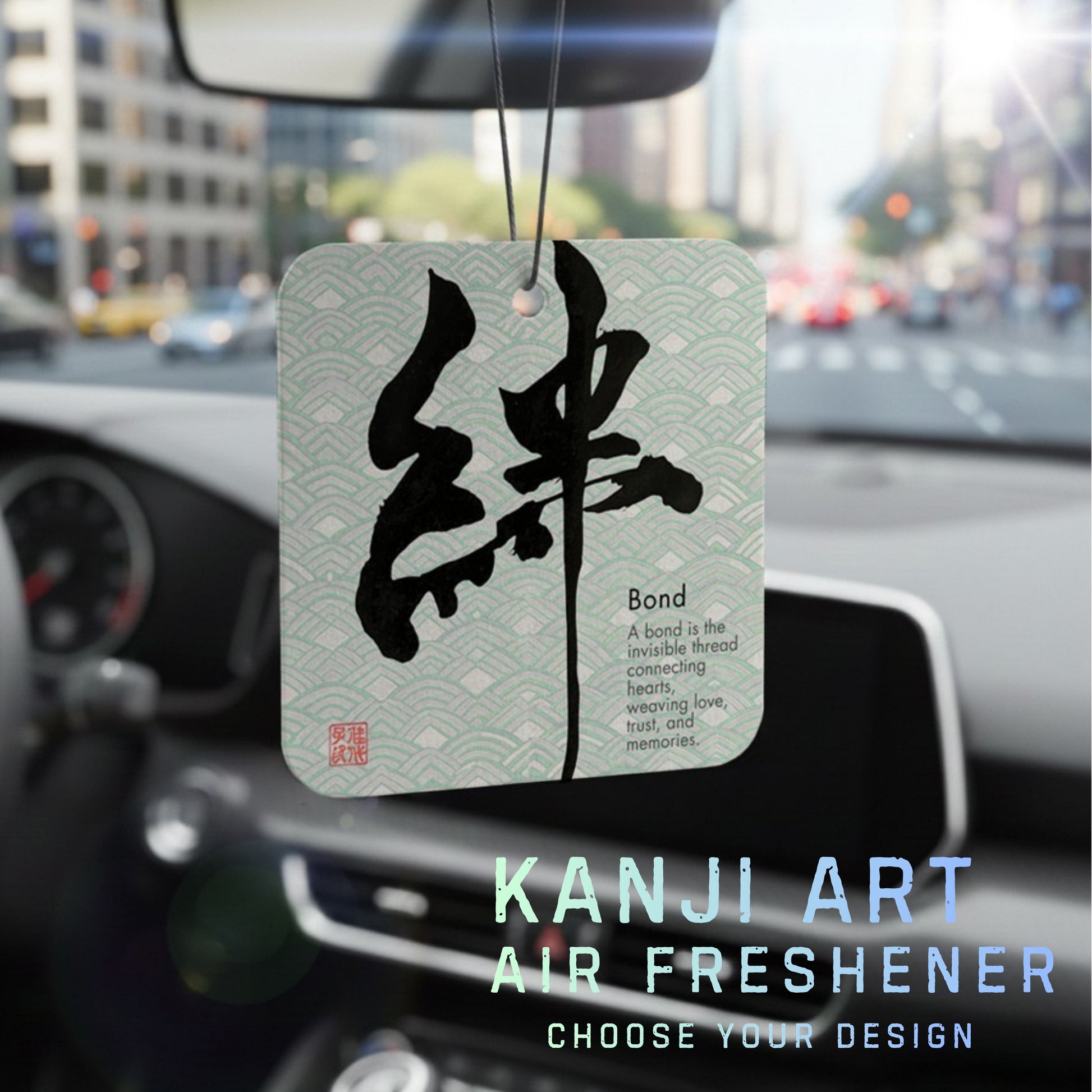 Kanji art air freshener hanging from a car's rearview mirror with a blurred cityscape background.