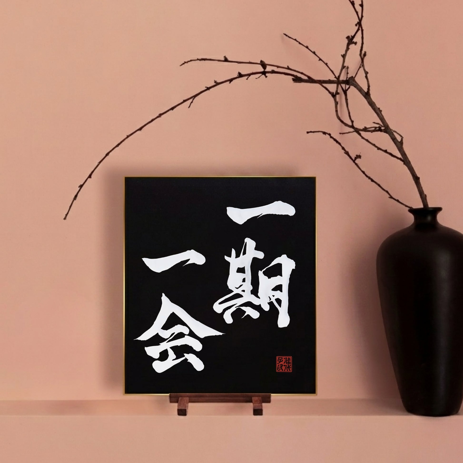 Framed artwork with white Japanese calligraphy on a black background, placed on an easel against a pink wall with a black vase and branch.