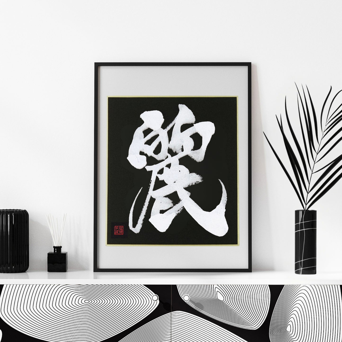 A high-contrast Japanese calligraphy print of 'Rei' (Beautiful/Graceful) on a black background with gold trim, in a black frame. Set in a modern Japandi interior with black vases and diffusers. Perfect wall art for a sophisticated living space.